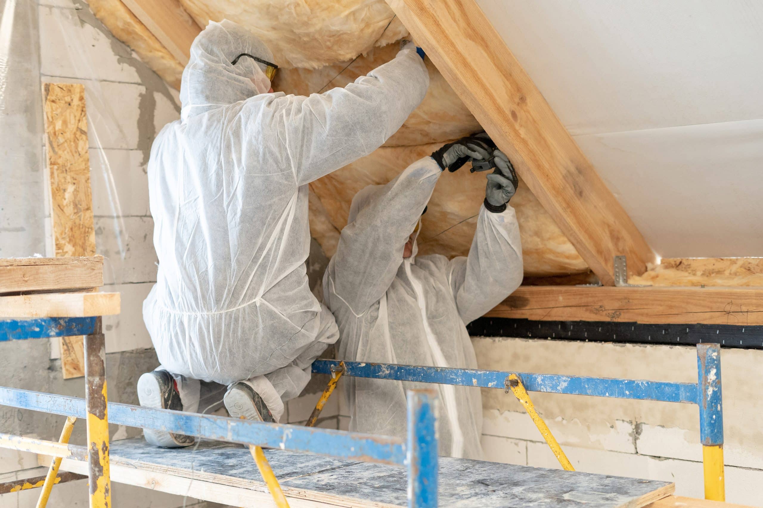Deux personnes en combinaisons de protection blanches installent une isolation sous un toit en bois incliné à l'intérieur d'un bâtiment inachevé, debout sur un échafaudage, travaillant soigneusement sur l'isolation maison.